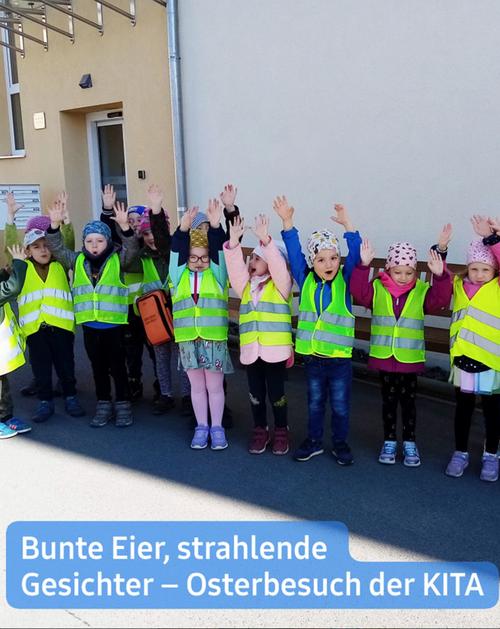 Eine Gruppe von Kindern, überwiegend im Vorschulalter, steht draußen an einer hellen Gebäudeoberfläche aufgereiht. Alle tragen leuchtend gelbe Sicherheitswesten und haben ihre Arme jubelnd in die Höhe gestreckt. Einige der Kinder sind mit Hüten oder Haarbändern geschmückt. Ihre Gesichter strahlen vor Freude und Ausdruckskraft.  Der Hintergrund zeigt ein schlichtes Gebäude mit einer hellen Fassade sowie sichtbaren architektonischen Details wie Fenstern und einer Metallkonstruktion in der Nähe des Daches. Der Boden scheint gepflastert zu sein.  Am unteren Rand des Bildes präsentiert ein hellblaues Banner einen Text, der die Szene eines Osterbesuchs ("Osterbesuch") in einer Kindertagesstätte ("Kita") beschreibt, inklusive farbenfroher Eier ("bunte Eier") und strahlender Gesichter ("strahlende Gesichter").