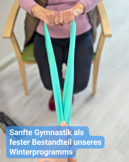Eine ältere Person sitzt auf einem Stuhl und führt eine Übung mit einem türkisfarbenen Theraband aus. Beide Hände halten das Band gespannt nach vorne, die Bewegung ist ruhig und kontrolliert. Die Szene findet in einem hellen Raum eines Pflegeheims statt und zeigt eine sanfte Gymnastikübung als Teil des Winterprogramms.